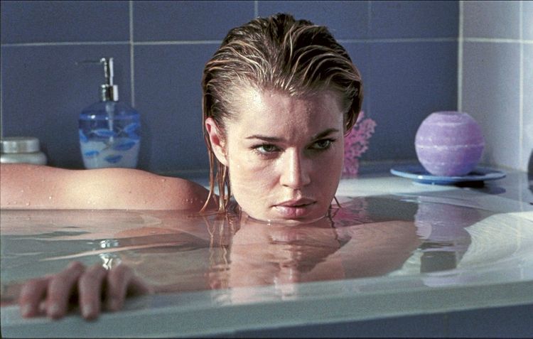 Eine Frau mit nassem Haar liegt in einer Badewanne, wobei nur ihr Gesicht und eine Hand sichtbar sind. Ihr Ausdruck wirkt ernst und nachdenklich. Im Hintergrund sind Badzubehör und Kacheln zu sehen.