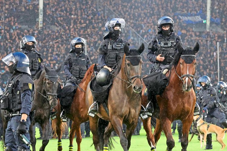 Polizeikräfte auf Pferden und zu Fuß, ausgestattet mit Schutzkleidung und Helmen, greifen auf einem Fußballfeld ein. Im Hintergrund sind schemenhaft Zuschauer zu erkennen.