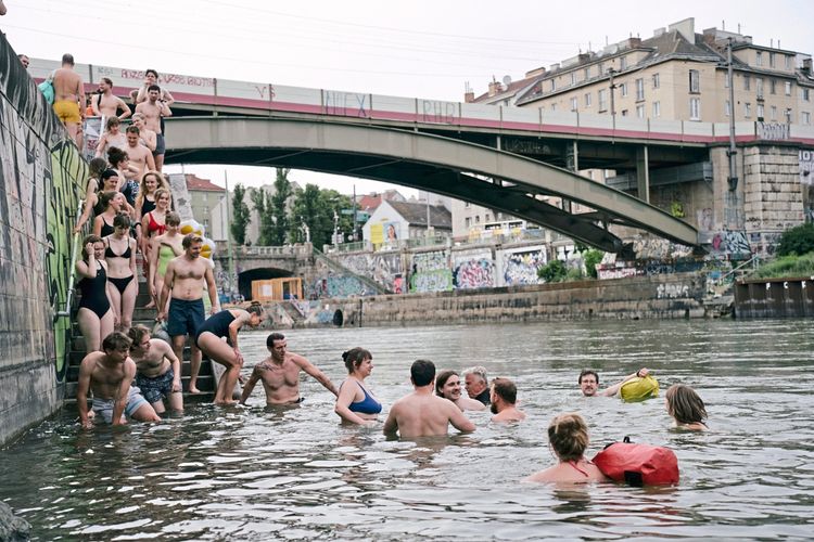 Eine Gruppe von Menschen badet und steht im Wasser eines Flusses. Einige steigen eine Treppe hinab, die mit Graffiti bemalt ist. Im Hintergrund ist eine Brücke sowie mit Graffiti versehene Wände und städtische Gebäude zu sehen.