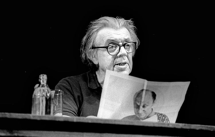 Schwarz-Weiß-Foto von einem älteren Mann mit Brille, der an einem Tisch sitzt und ein Blatt Papier mit einem Bild darauf hält. Neben ihm stehen eine Karaffe und ein Glas. Aufnahme aus einer Lesung im Deutschen Theater in Berlin, 16.10.1988.