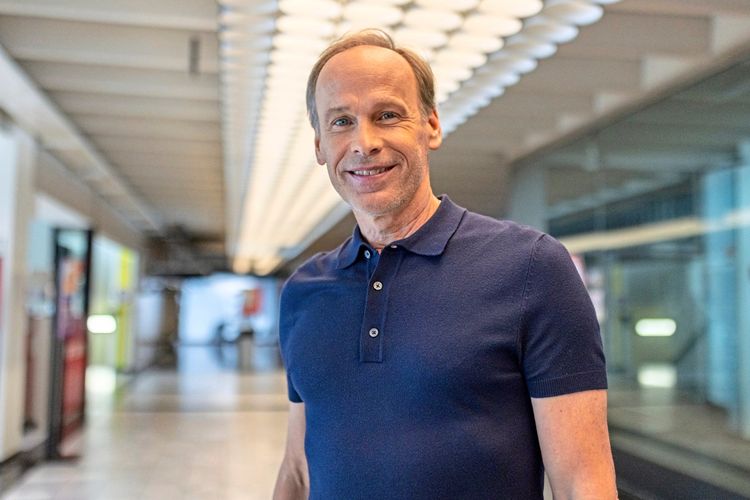 Ein Mann in einem blauen Poloshirt steht in einem modern gestalteten Innenbereich mit Glaswänden und Deckenleuchten. Er lächelt in die Kamera.