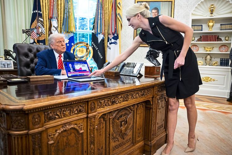 US-Präsident Donald Trump sitzt an seinem Schreibtisch im Oval Office und beobachtet, wie eine Frau in einem schwarzen Kleid einen Laptop mit einer Nachricht über die Börse auf den Schreibtisch stellt. Im Hintergrund sind Fahnen und Bücherregale zu sehen.
