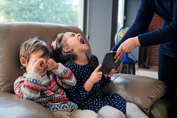 Eine Frau nimmt einem weinenden Mädchen auf einem Sofa ein Tablet weg, während ein Junge daneben sitzt und sich die Augen reibt. Thema: Abhängigkeit von digitalen Geräten.