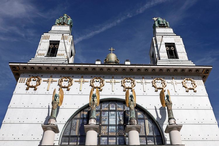 Ansicht der Kirche am Steinhof in Wien, Österreich. Das Gebäude, im Jugendstil von Otto Wagner entworfen, zeigt detailreiche Skulpturen von Engeln, gestaltet von Othmar Schimkowitz. Goldene Verzierungen und Symbole schmücken die weiße Fassade unter einem klaren blauen Himmel.