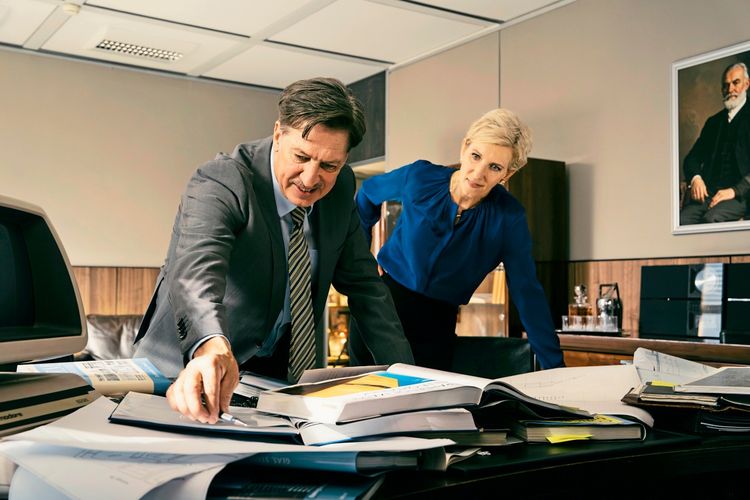 Ein Mann im Anzug und eine Frau in einer blauen Bluse stehen in einem Büro und beugen sich über einen vollgepackten Schreibtisch mit Dokumenten und Unterlagen. Im Hintergrund ist ein Gemälde an der Wand sichtbar.