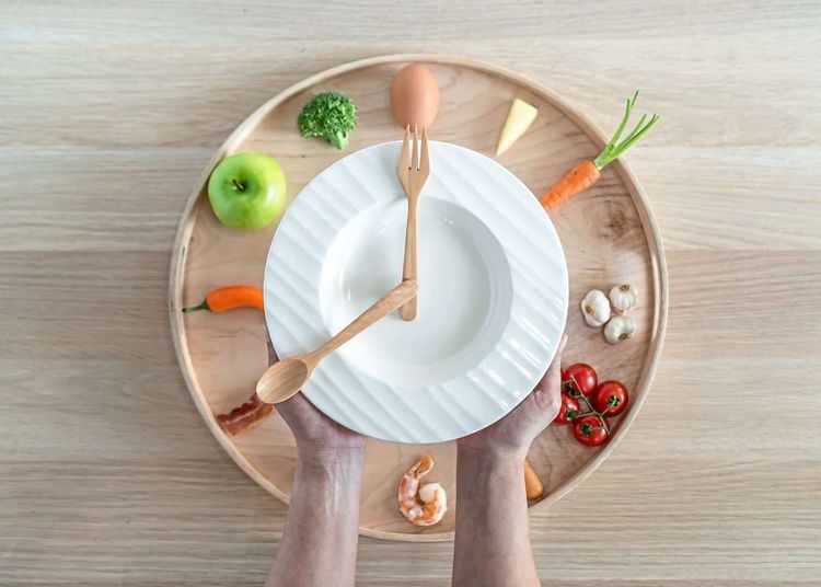 Zwei Hände halten einen leeren Teller, auf dem Holzbesteck acht Uhr anzeigt, über Gemüse
