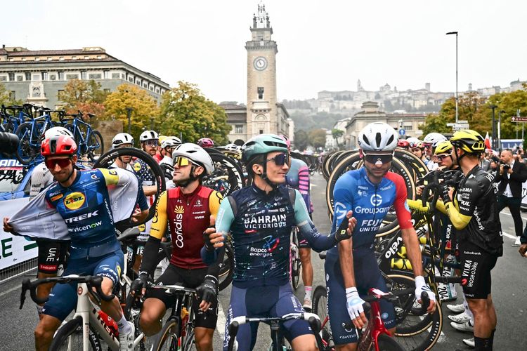 Radrennfahrer stehen vor dem Start der 120. Ausgabe des Giro di Lombardia in Bergamo. Im Vordergrund sind mehrere Fahrer in Teamtrikots zu sehen, darunter ein Fahrer des Bahrain Victorious-Teams, der gestikuliert. Im Hintergrund ist die Skyline von Bergamo mit dem Turm eines historischen Gebäudes sichtbar. Fahrräder und weitere Teilnehmer sind ebenfalls im Bild.
