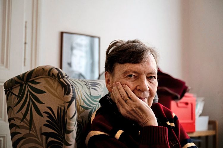 Ein älterer Mann sitzt in einem Sessel mit gemustertem Bezug und stützt seinen Kopf nachdenklich mit der Hand. Im Hintergrund ist ein unscharfes Schwarz-Weiß-Foto an der Wand sowie ein roter Gegenstand zu sehen.
