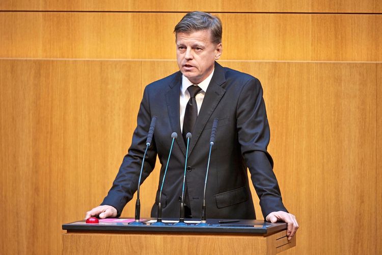 Ein Mann im schwarzen Anzug und schwarzer Krawatte steht am Rednerpult mit mehreren Mikrofonen in einem hölzernen Saal.