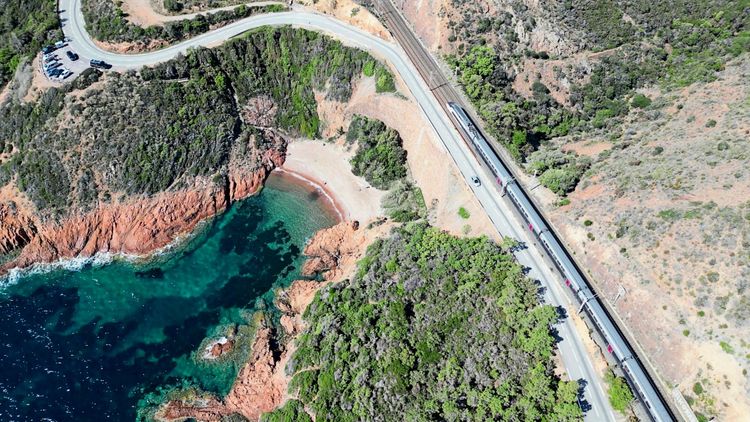 Luftaufnahme einer Küstenlandschaft mit klarem türkisfarbenem Wasser, einer kleinen Bucht, umgeben von roten Klippen und grüner Vegetation. Rechts verläuft eine Straße parallel zu einer Bahnschiene, auf der ein Zug fährt. Links oben befinden sich geparkte Autos auf einem Parkplatz.