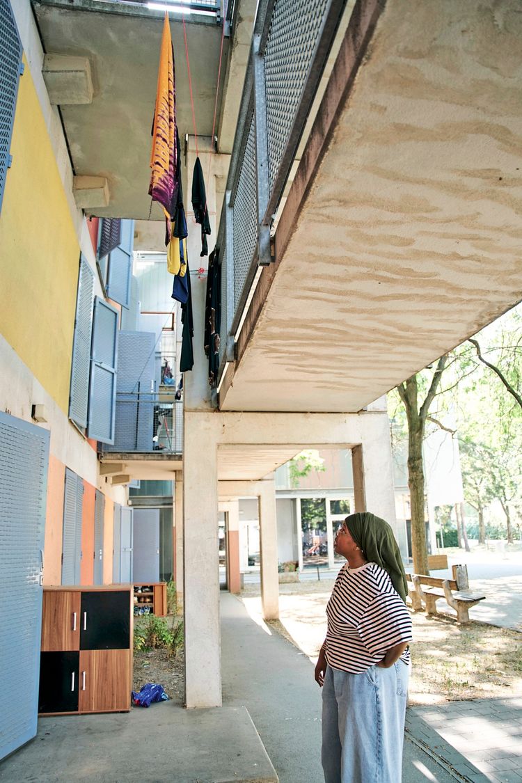 Eine Person steht draußen in der Macondo-Siedlung in Wien, Simmering, und blickt nach oben zu einer Wäscheleine mit aufgehängter Kleidung. Die Umgebung zeigt Wohngebäude, Balkone und Grünbereiche.