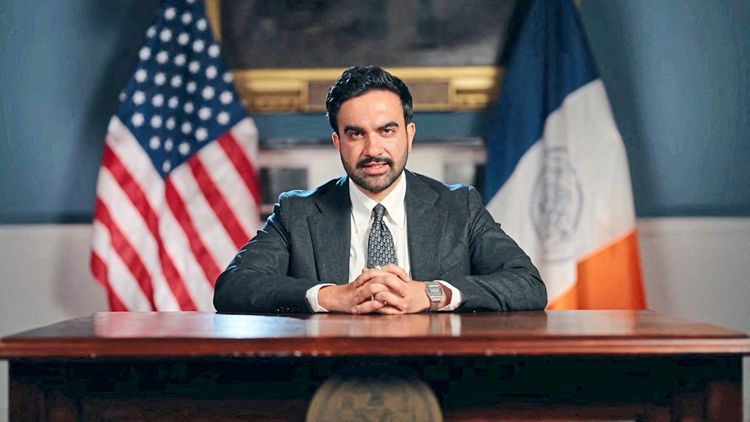 Ein Mann in einem Anzug sitzt an einem hölzernen Schreibtisch, die Hände gefaltet, mit einer US-amerikanischen Flagge und der Flagge von New York City im Hintergrund.