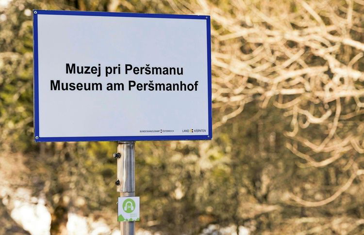Ein Schild mit der Aufschrift „Muzej pri Peršmanu / Museum am Peršmanhof“ in Deutsch und Slowenisch, aufgenommen in einer bewaldeten Umgebung in Bad Eisenkappel, Kärnten.