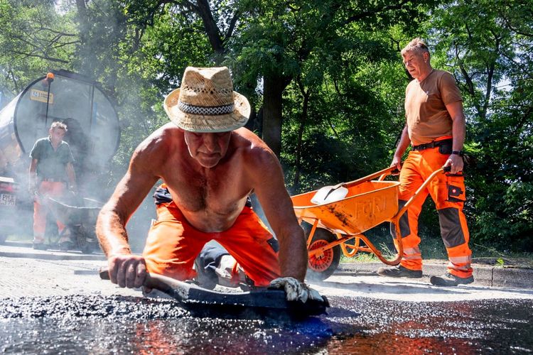 Eine Baustellenszene bei sommerlicher Hitze: Ein Arbeiter mit Strohhut und freiem Oberkörper glättet bei 33 Grad heißem Wetter Gussasphalt auf einer Straße. Im Hintergrund sind zwei weitere Arbeiter mit Schubkarre und Arbeitsmaterialien zu sehen.