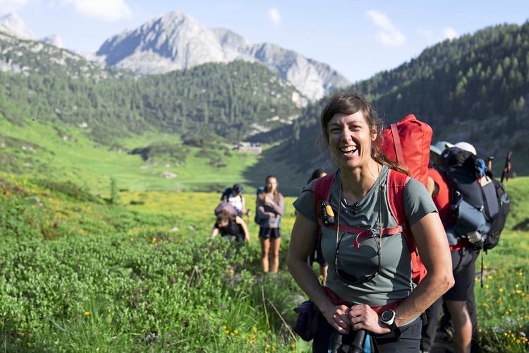 Eine lachende Frau mit rotem Rucksack steht auf einer Bergwiese in einer alpinen Landschaft. Im Hintergrund sind weitere Wanderer, grüne Hügel und felsige Berge zu sehen.