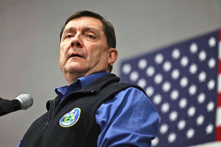 Ein Mann in einem blauen Hemd und einer schwarzen Weste spricht in ein Mikrofon. Im Hintergrund ist eine amerikanische Flagge zu sehen.
