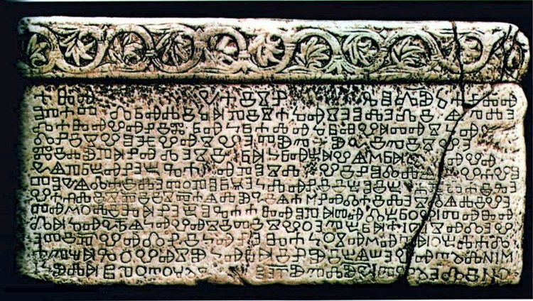 Steintafel mit Gravuren in einer antiken Schrift, übersät mit Reihen unbekannter Schriftzeichen. Am oberen Rand befindet sich ein dekoratives Muster aus floralen Motiven. Die Tafel weist altersbedingte Risse und Verfärbungen auf.