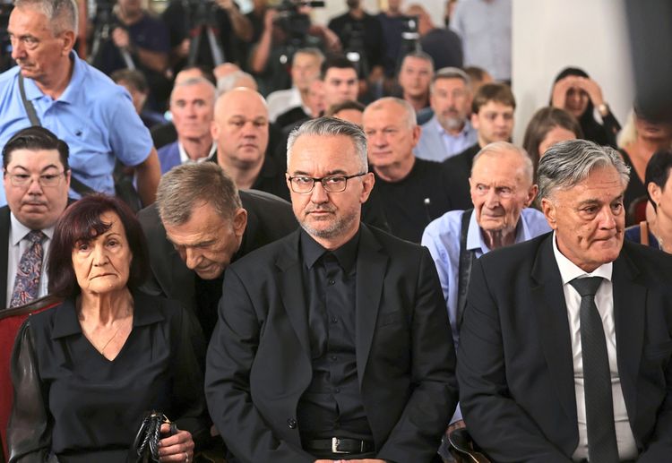 Eine Gruppe von Menschen sitzt in einem Veranstaltungsraum, darunter eine ältere Frau links, ein Mann in der Mitte mit Brille und dunklem Anzug sowie ein älterer Mann rechts in schwarzem Anzug. Die Stimmung im Raum wirkt ernst und andächtig. Im Hintergrund sitzen weitere Personen und einige Kameras sind aufgestellt.