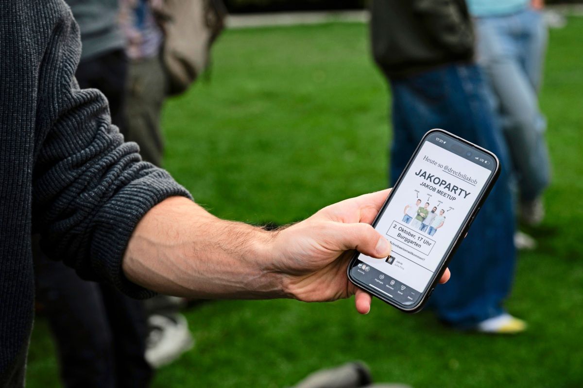 Ein Arm hält ein Smartphone, auf dessen Bildschirm eine Einladung zu einer „JAKOPARTY – Jakob Meetup“ am 2. Oktober im Burggarten angezeigt wird. Im Hintergrund sind unscharf stehende Personen und eine Rasenfläche zu sehen.