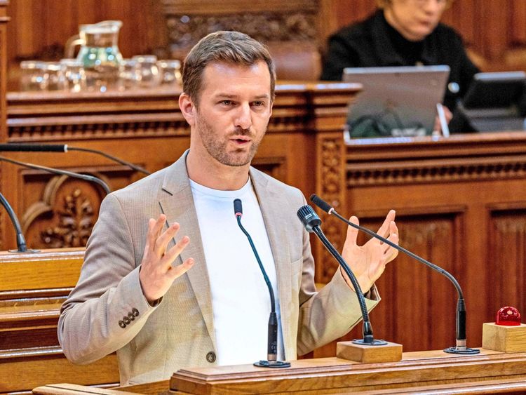 Ein Mann steht an einem Rednerpult im Wiener Rathaus und hält eine Rede. Er trägt ein hellbeiges Sakko und ein weißes T-Shirt, gestikuliert mit beiden Händen und spricht in Mikrofone. Im Hintergrund sind Holzelemente und eine weitere Person am Laptop zu sehen.