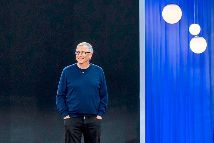 Ein Mann mittleren Alters mit Brille und dunkelblauem Pullover steht lächelnd vor einem dunklen Hintergrund. Rechts im Bild hängen runde Lampen vor einem blauen Vorhang.