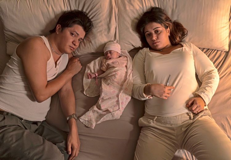 Eine Frau und ein Mann liegen auf einem Bett, mit einem Neugeborenen dazwischen. Das Baby ist in eine rosa Decke gehüllt und trägt eine Mütze. Die Stimmung wirkt nachdenklich.
