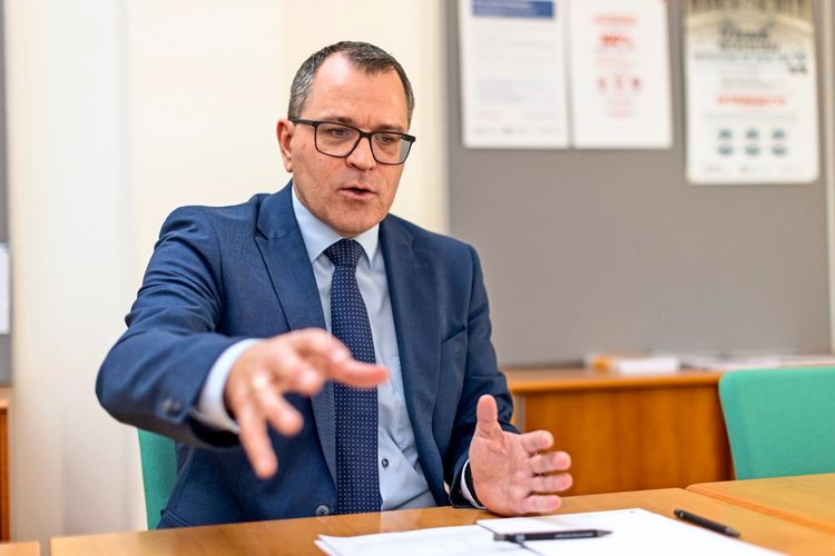 Gernot Kanduth in einem blauen Anzug und mit Brille sitzt an einem Tisch und gestikuliert mit der rechten Hand. Auf dem Tisch liegen Papierdokumente und ein Kugelschreiber. Im Hintergrund sind Poster und ein Regal erkennbar.