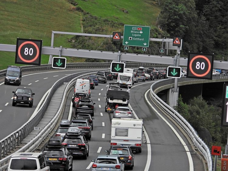Stau auf der Autobahn vor dem Nordportal des Gotthardtunnels in der Schweiz. Verkehrsschilder zeigen eine Geschwindigkeitsbegrenzung von 80 km/h und grüne Pfeile für geöffnete Spuren. Sommerliche Landschaft im Hintergrund.