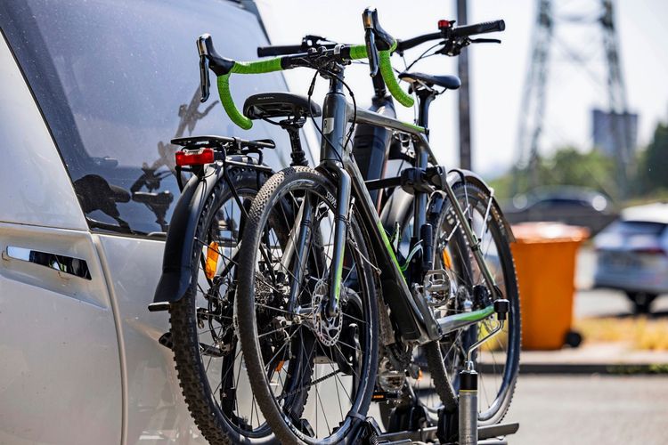 Fahrräder auf einem Fahrradträger, der an einem silbernen Wohnwagen befestigt ist. Im Hintergrund unscharf weitere Fahrzeuge, ein Strommast und eine orangefarbene Mülltonne.