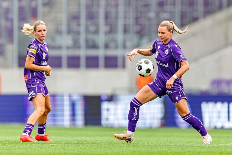 Zwei Fußballspielerinnen des FK Austria Wien Frauen-Teams in lila Trikots während eines Spiels in der Generali Arena. Eine Spielerin balanciert den Ball, die andere schaut aufmerksam zu. Im Hintergrund sind unscharfe Tribünen zu sehen.