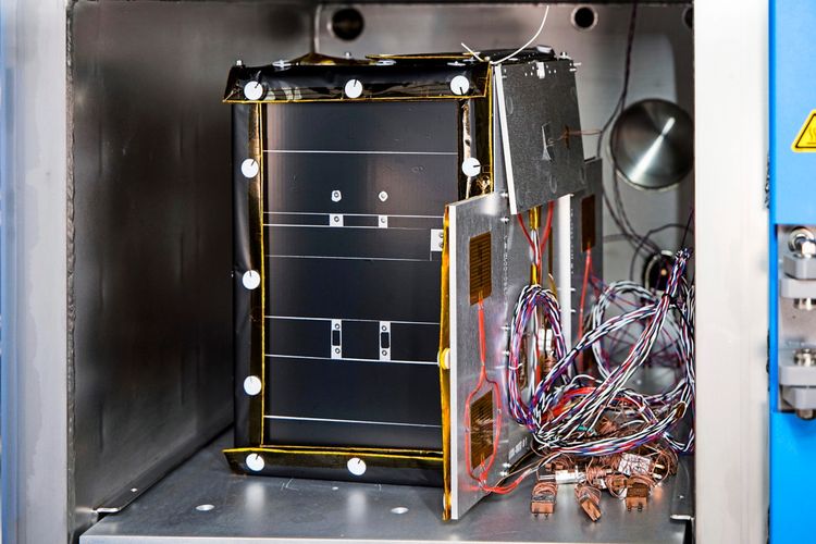 Ein technisches Gerät, vermutlich ein Satellitenmodell, wird in einer Thermalkammer getestet. Das Gerät ist mit Kabeln und Drähten verbunden, die rechts aus dem Gehäuse herausragen. Die Umgebung deutet auf ein Labor oder eine Testeinrichtung hin.