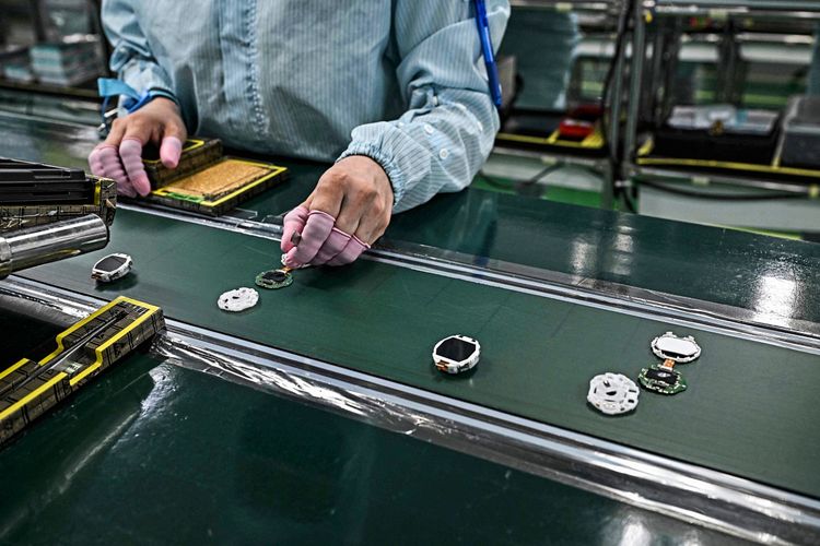 Eine Person in Schutzkleidung und mit rosa Fingerlingen montiert Bauteile von Uhren an einem Fließband in einer Fabrik. Elektronische Komponenten und Uhrengehäuse sind auf der Arbeitsfläche zu sehen.