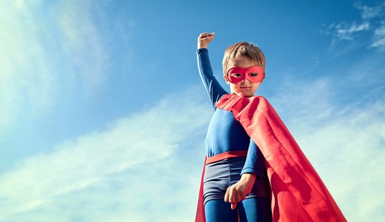 Ein Kind in rotem Cape und mit roter Augenmaske posiert vor blauem Himmel in Superheldenpose, eine Faust in der Luft.