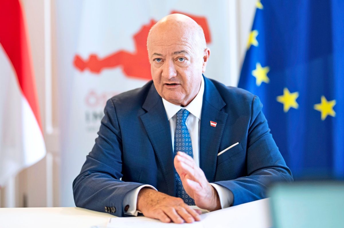 Ein Mann in einem Anzug sitzt an einem Tisch und gestikuliert, während er spricht. Im Hintergrund sind eine Karte von Österreich sowie die Flaggen Österreichs und der Europäischen Union sichtbar.