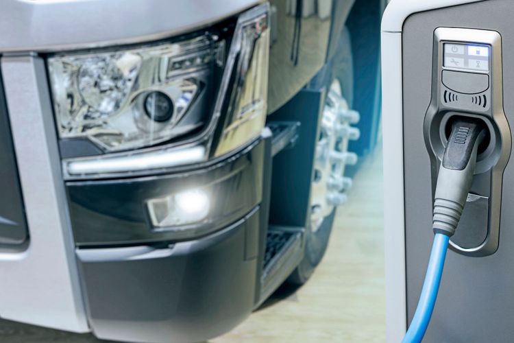 Ein Elektro-Lkw wird an einer Ladestation mit einem blauen Ladekabel aufgeladen. Im Vordergrund ist der Ladeanschluss zu sehen, im Hintergrund der Frontbereich des Lkw mit Scheinwerferdetails.