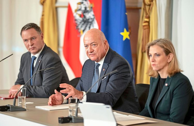 Drei Personen sitzen an einem Tisch während einer Pressekonferenz im Bundeskanzleramt in Wien. Im Hintergrund sind die österreichische und die EU-Flagge zu sehen.