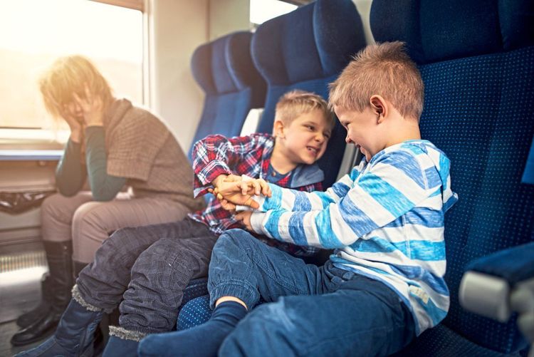 Zwei streitende Kinder im Zug, eine verzweifelt wirkende Mutter sitzt daneben.