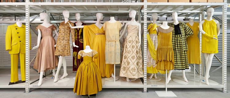 Schaufensterpuppen in einem Museumslager, gekleidet in Kleidungsstücke verschiedener Stile und Gelb- sowie Beigetöne. Die Kleidung umfasst Kleider, Anzüge und Ensembles aus unterschiedlichen Epochen, die auf weißen Regalen präsentiert werden.