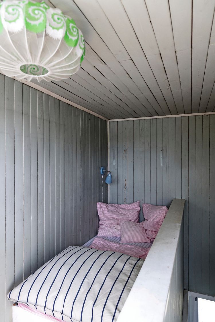 Ein schlichtes Schlafzimmer mit Holzvertäfelung in hellem Grau. Das Bett ist mit weißer Bettwäsche mit blauen Streifen und rosa karierten Kissen ausgestattet. Eine blaue Wandleuchte und eine weiße Papierlampe mit grünem Muster hängen an der Wand und Decke.